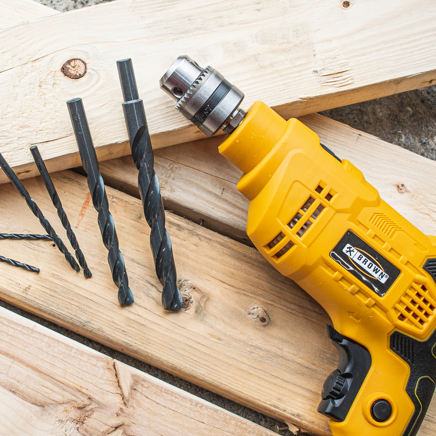 Yellow Brown brand power drill with three black drill bits on wooden boards, highlighting tools in a woodworking setup.